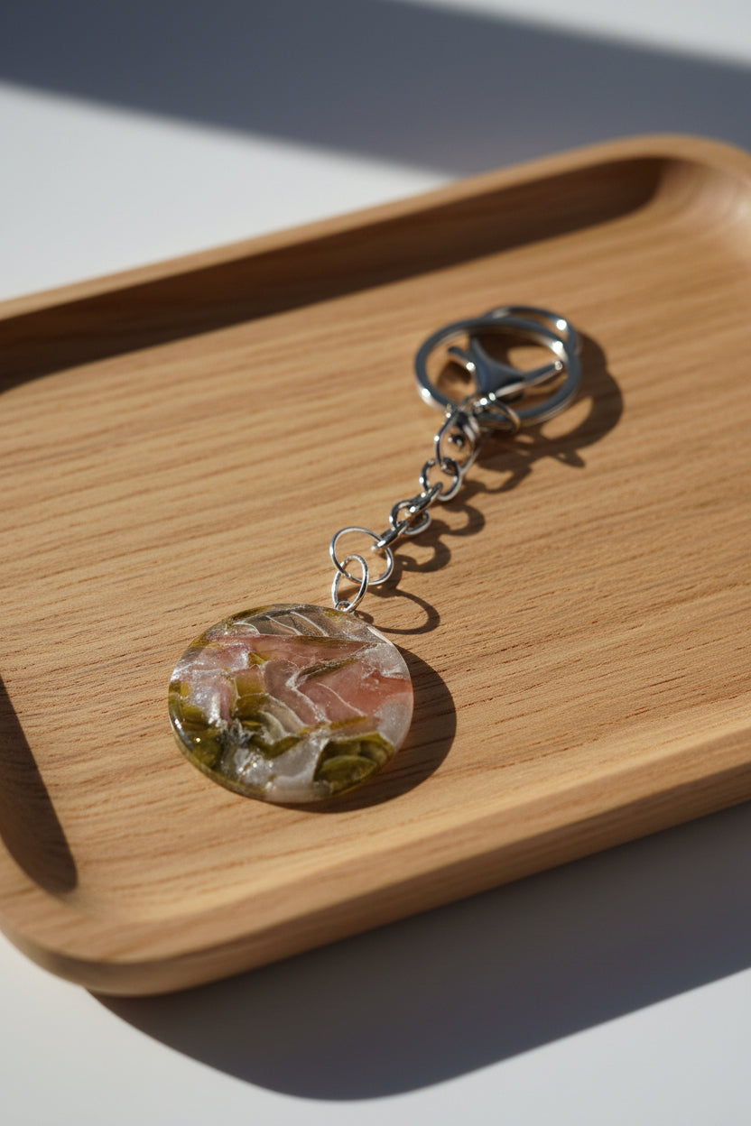 A round resin keychain charm with a marbled pattern of pink, green, and gold, attached to a silver metal split ring and chain, photographed on a white background with a hard shadow.