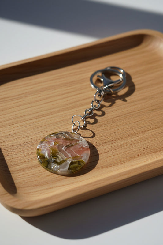 A round resin keychain charm with a marbled pattern of pink, green, and gold, attached to a silver metal split ring and chain, photographed on a white background with a hard shadow.